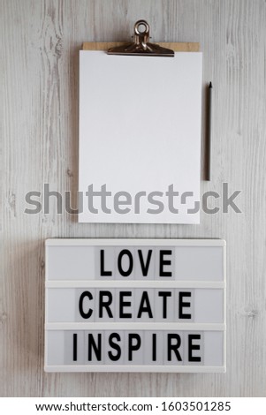 'Love create inspire' words on a modern board, clipboard with blank sheet of paper on a white wooden background, top view. Overhead, from above, flat lay. 