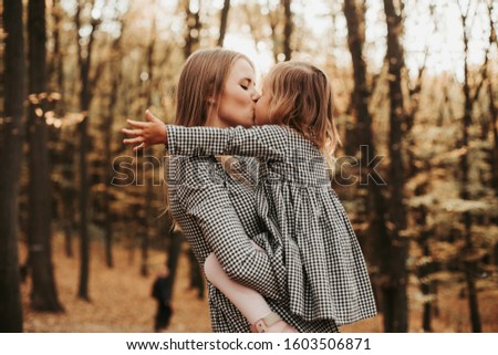 Beautiful amazing and lovely picture of young mother kissing her daughter. Stand together in autumn park or forest. Woman hold girl in hands. Tender picture. Unconditional love. Autumn sunset
