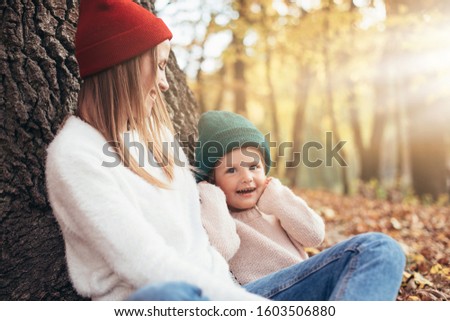 Picture of young woman and child in colorful hats and same color sweaters with jeans sitting together and posing. Side view. Daughter look on caeram and smile. In autumn forest or park