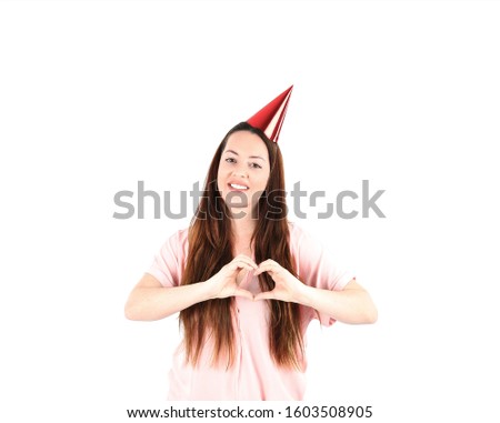 Cute young woman smiling and doing a heart symbol with both hands while wearing a red party hat against a white background