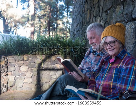Attractive elderly couple are resting reading a book outdoors. Focus on the man's face. Casual dresses with colored and checkered shirts. Sitting in a sheltered corner of stones