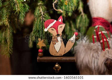 Cute handmade Christmas tree decoration in shape of nice and funny wooden fox in Santas hat and cozy scarf. Handcrafted new year 2021 toy with background of green Christmas tree lights garland