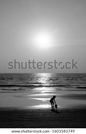 Silhouette of an unrecognizable small child holding a football walking along an empty golden sandy beach at sunset, a calm sea and clear golden sun lit sky, black and white photograph 