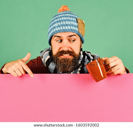 Autumn and cold weather concept. October beverage idea. Hipster with beard and scared face looks at tea or coffee cup pointing down. Man in hat holds brown cup on green and pink background, copy space