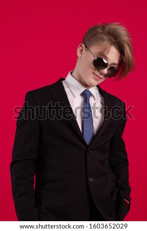 Young teenager guy in a black jacket, tie and sunglasses posing on a red background