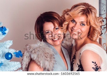 Elegant girlfriends in evening dresses and a fur collar stand nearby. Sisters hug. Young beautiful women smiling in anticipation of Christmas, gathering to visit friends. 