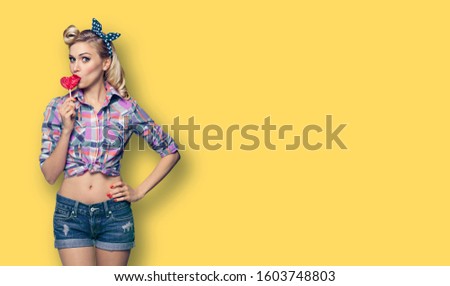 Surprised woman eating heart shape lollipop. Girl in pin up cloth. Retro fashion and vintage concept picture. Yellow color background. Copy space for some advertise slogan or text. 