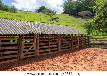 A kind of barn on a corral, after a heavy rain with the ground full of puddles. In the background there are a green pasture and blue sky