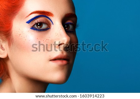 Beautiful girl with colored in red hair and blue eyeliner makeup isolated on blue background Royalty-Free Stock Photo #1603914223