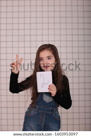 young smiling girl of eight years with long hair in a black turtleneck and denim sundress with a sign "2 two"