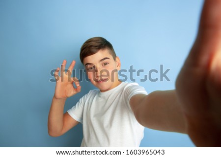 Taking selfie with sign of nice. Caucasian boy portrait isolated on blue studio background. Beautiful teen male model in white shirt posing. Concept of human emotions, facial expression, sales, ad.