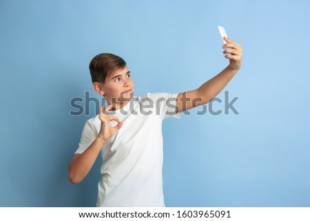 Taking selfie or rolling vlog. Caucasian boy portrait isolated on blue studio background. Beautiful teen male model in white shirt posing. Concept of human emotions, facial expression, sales, ad.