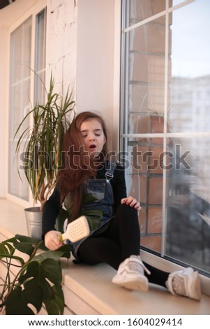 young smiling girl of eight years with long hair in a black turtleneck and jeans sundress sits on a window sill by the window and combs her hair with a comb