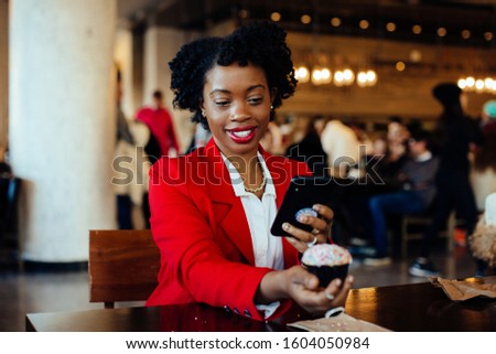 Portrait of a smiling young woman sitting in a cafe taking a cell phone picture of her cupcake