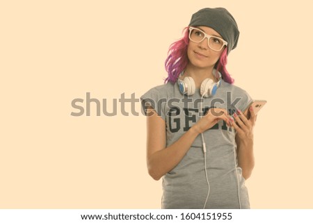 Studio shot of geek girl holding mobile phone while thinking with headphones around neck