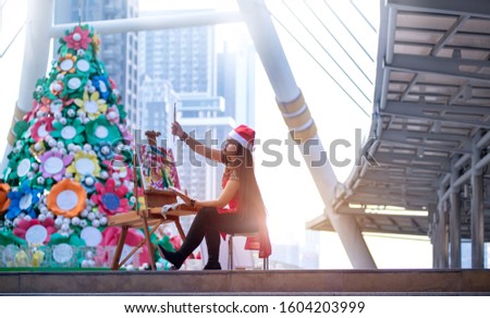 Street female artist Painting abstract art in the modern city, with Santa hat on head, big Christmas tree in background