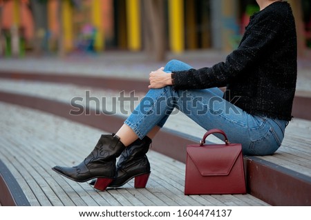 Fashionable young woman in high heel black cowboy boots, blue jeans, black tweed jacket and burgundy handbag on the city street. Street style. Royalty-Free Stock Photo #1604474137