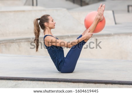 Pilates model with a thin athletic body and beautiful face, wearing dark blue clothes, holding a V pose with a small orange Pilates ball between her legs. Urban skate park background.