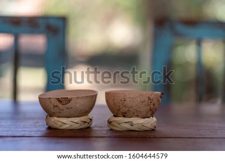 Two traditional "jicama" cups for drinking mezcal in Mexico, on their "rodete" stands. Cups made from the skin of the gourd fruit of a jicama tree. On an outdoor table with two rustic chairs behind. Royalty-Free Stock Photo #1604644579