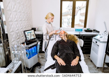 Blonde female beautician doctor applying red led light therapy to female client in cosmetology clinic, facial photo therapy. Anti-aging treatments and photo rejuvenation procedure