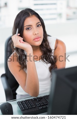 Thoughtful cute businesswoman holding pen looking at camera in bright office
