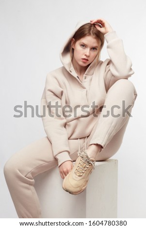 Young girl wearing blank and oversize beige long hoody, beige pants, beige sneakers . Sitting on white cube. White background.
