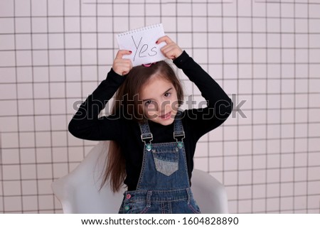 young kind playful girl of eight years with long hair in a black turtleneck and denim sundress with the sign "YES"