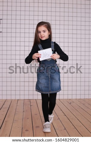 young kind playful girl of eight years with long hair in a black turtleneck and denim sundress with the sign "YES"