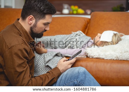 Bearded man browsing medical sites on smartphone while sick little girl lying on couch stock photo