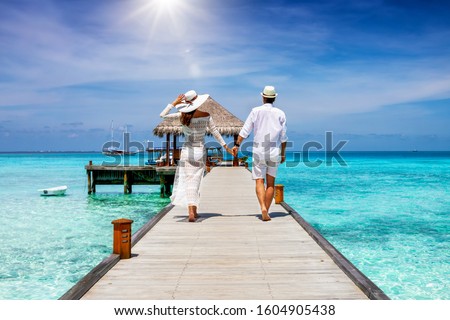A happy couple in white summer clothing on vacation walks along a wooden pier over tropical, turquoise ocean in the Maldives, Indian Ocean Royalty-Free Stock Photo #1604905438