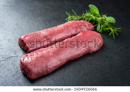 Raw dry aged venison tenderloin fillet steak natural with herbs offered as closeup on a modern design board  Royalty-Free Stock Photo #1604926066