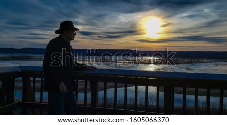 Older man in hat looking at sunset over Missouri River flood plane from observation deck; dark blue sky in background; sun reflects in water 