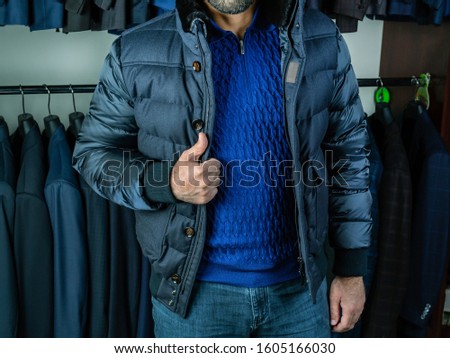 A young man with a blue sweater and jeans demonstrates a winter jacket. Shades of blue. Outerwear in a boutique