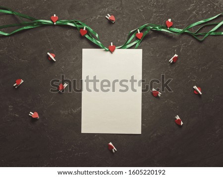 A blank sheet of paper is on a dark texture background. Nearby are a decorative green ribbon and wooden clothespins with red hearts. Composition for Valentine's Day. Copyspace. Flat lay.