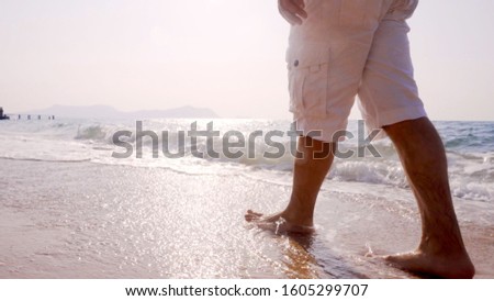 A man is walking along the golden sand of the beach. Male legs walking near the sea. Bare feet of a guy walking along a sandy shore with waves. Summer vacation or vacation.