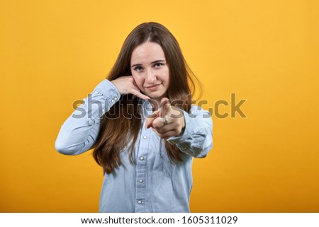 Attractive caucasian woman wearing fashion shirt isolated on orange background in studio doing call gesture on ear, pointing finger at camera. People emotions, lifestyle concept.