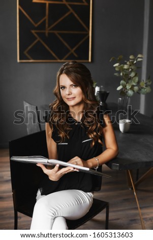 Young beautiful woman in black blouse and white pants reading an interesting book in the cafe interior. Happy model girl with trendy makeup and hairstyle sits at the table and reading a book