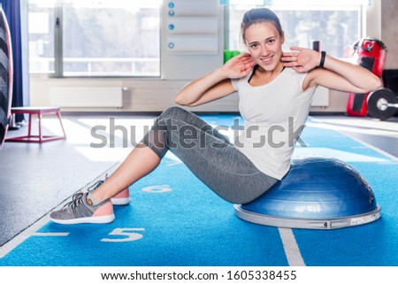 Focused sporty beautiful young athletic brunette woman in gray leggings and white t-shirt working in gym doing exercises for abdominal muscles on bosu balance trainer, lie down on fitness ball, indoor