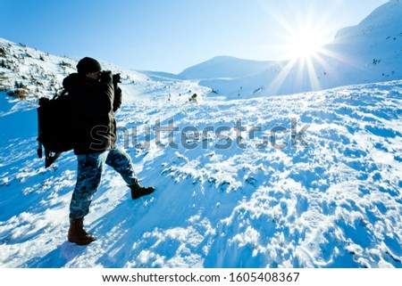 Young man photographer in winter clothing standing and making photo with camera in sunlight with white snow background. Travelling and making photos of winter nature concept