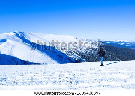 Young man photographer standing with camera on hill with panoramic view of winter valley, hills and forest on sunny clear winter day. Travelling and making photos of winter nature concept