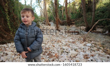 A boy with blond hair wearing a gray coat playing in a stream in a tree and shrub landscape and a stream flowing with stone soil