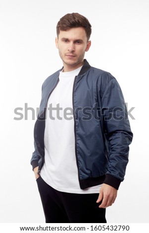 Young european man in white sweater and black pants, blue bomber jacket posing on white background. Isolated.