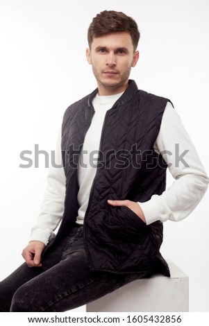 Handsome man in black vest, black jeans and boots isolated on white background. Sitting on white cube.