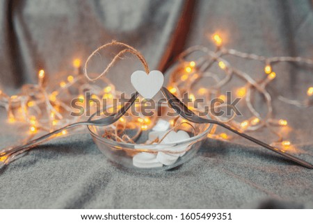 Saint Valentine's day card. White wooden hearts on the bright background. Hearts with garland lights. Spoon and fork hold a heart with bright bokeh.