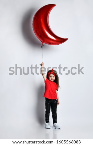 Small girl in red t-shirt, leather trousers, white sneakers and hair accessory standing and holding red moon shaped balloon in hand over white wall background. Stylish children clothing concept