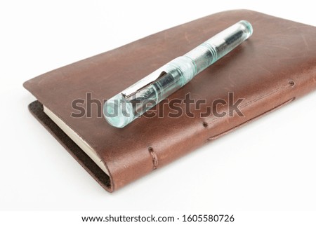 A simple but artful combination of classic leather bound journal and green glass fountain pen set on a plain white background.