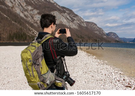 
A young man taking a photo with a smartphone. Tourist, traveler with a backpack, camera. A boy wearing a black jacket. A lake surrounded by mountains. Picturesque scenery. Beautiful landscape.