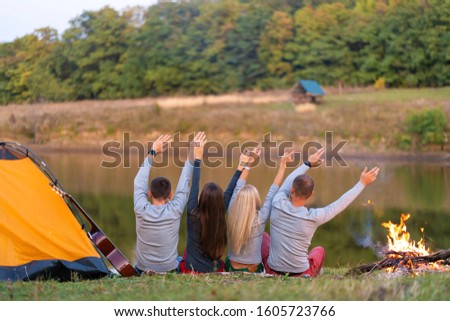 Shoot from back. A group of happy friends camping at riverside, dancing hold hands up  and enjoy view. Holidays fun.