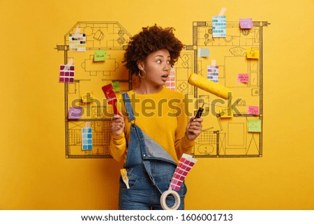 Photo of shocked curly woman plans home DIY renovation, holds paint tools, wears denim overalls, has color guide, poses indoor against house sketch, gives something lick, puts finishing touches