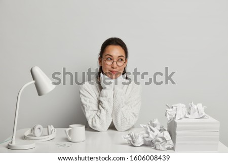 Pretty young Asian woman worker keeps hands under chin, looks aside with thoughtful expression, has nice plan in mind, wears round spectacles and sweater, has pile of blank white papers on table